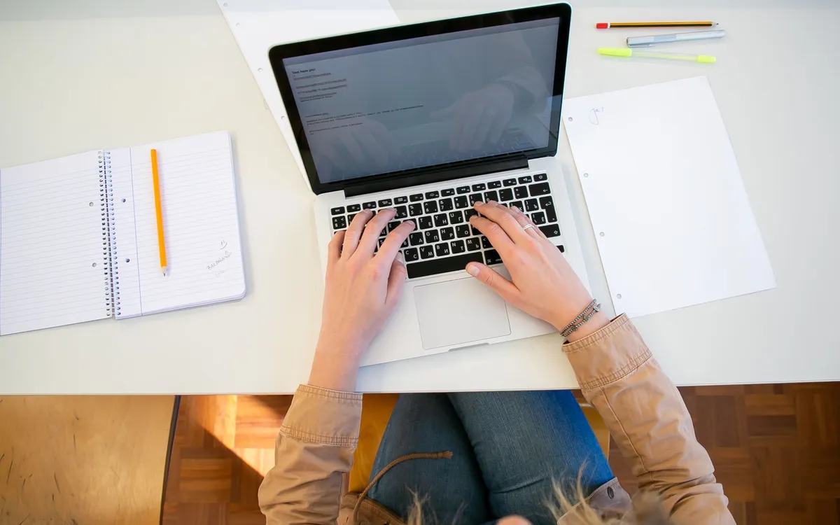 An overhead view of someone working at a laptop with notebooks and documents
